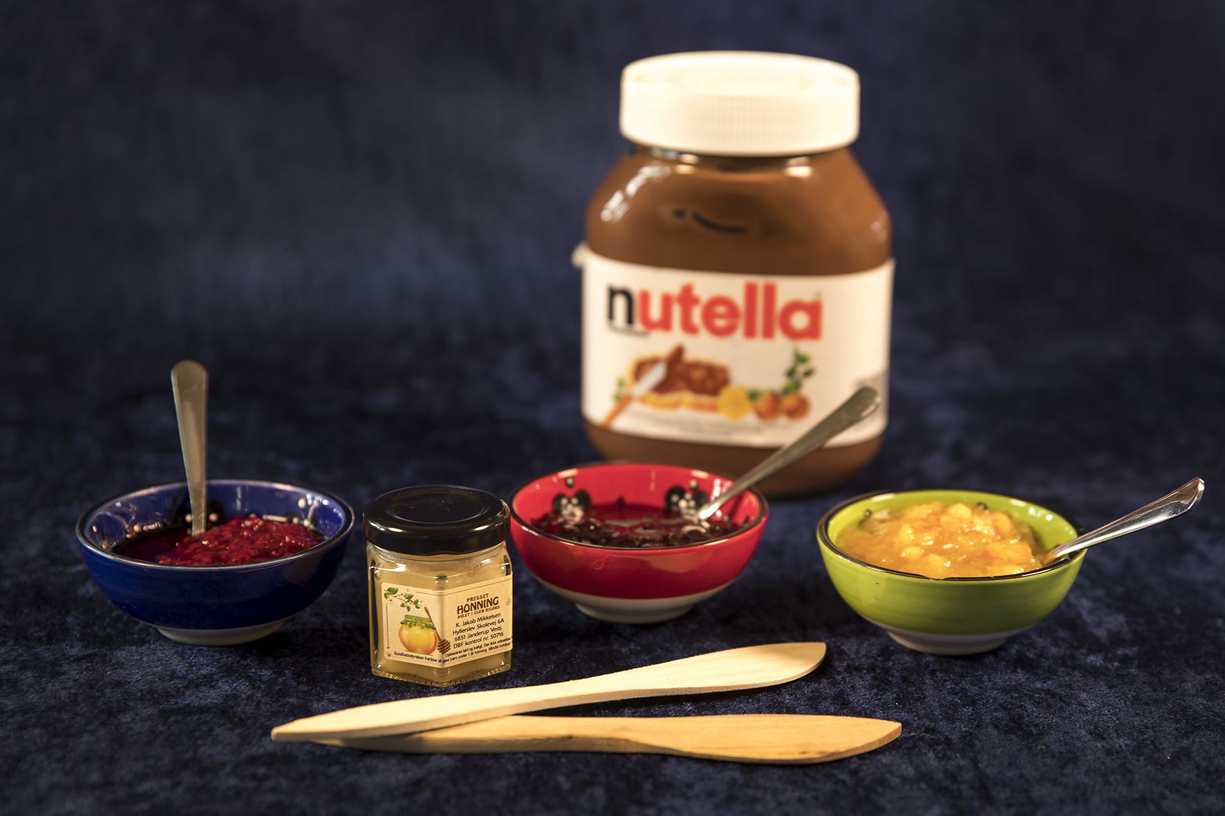 A Nutella jar, a jar of honey, and three bowls with different spreads on a dark surface.