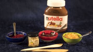 A Nutella jar, a jar of honey, and three bowls with different spreads on a dark surface.