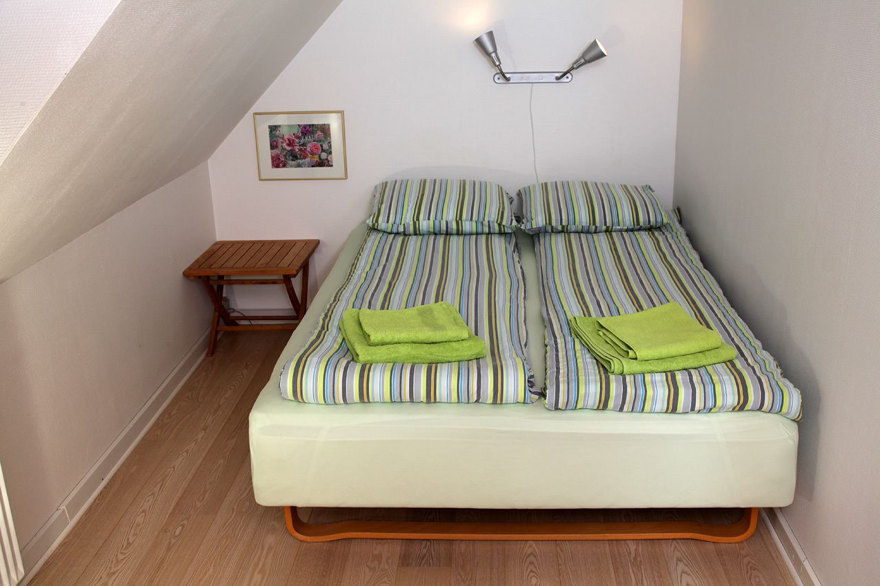 A small, neatly arranged bedroom with a single bed, a small wooden nightstand, a wall-mounted light, and a framed picture.