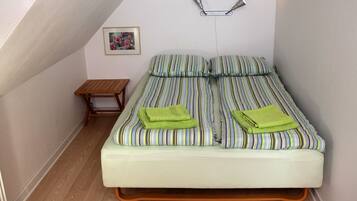 A small, neatly arranged bedroom with a single bed, a small wooden nightstand, a wall-mounted light, and a framed picture.