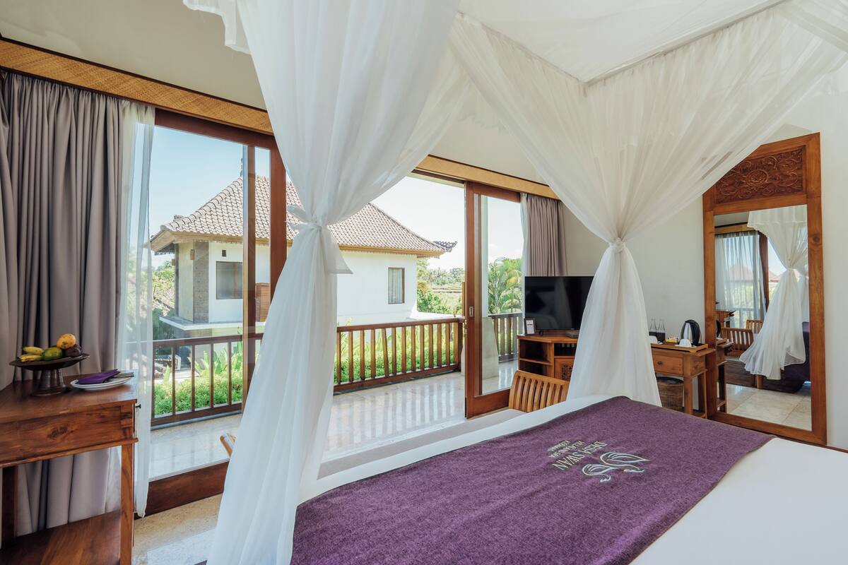 Une chambre avec un grand lit, une télévision, un bureau avec un ordinateur portable et un balcon donnant sur une maison.
