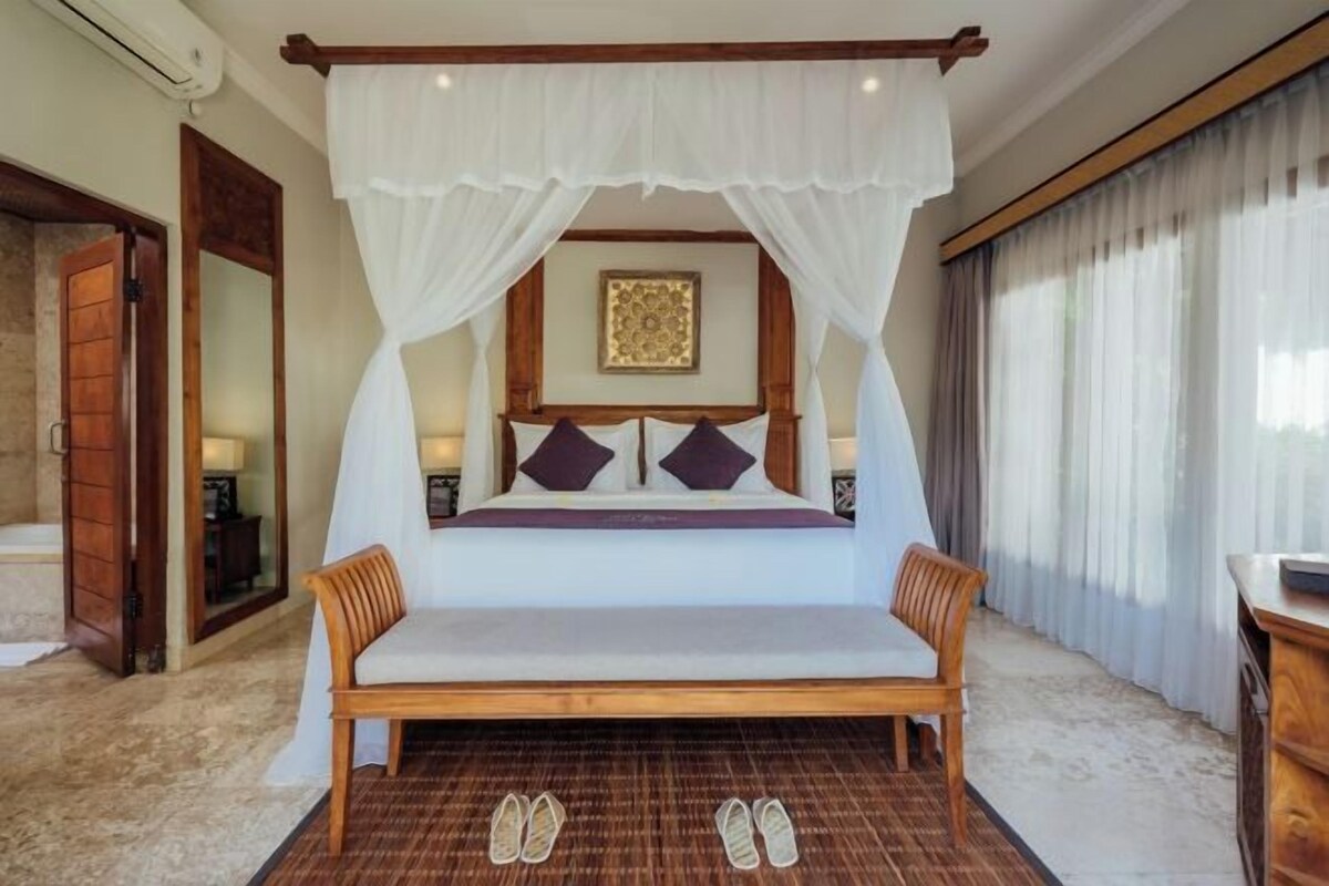 A bedroom with a canopy bed, a wooden bench, a mirror, and a large window with curtains.