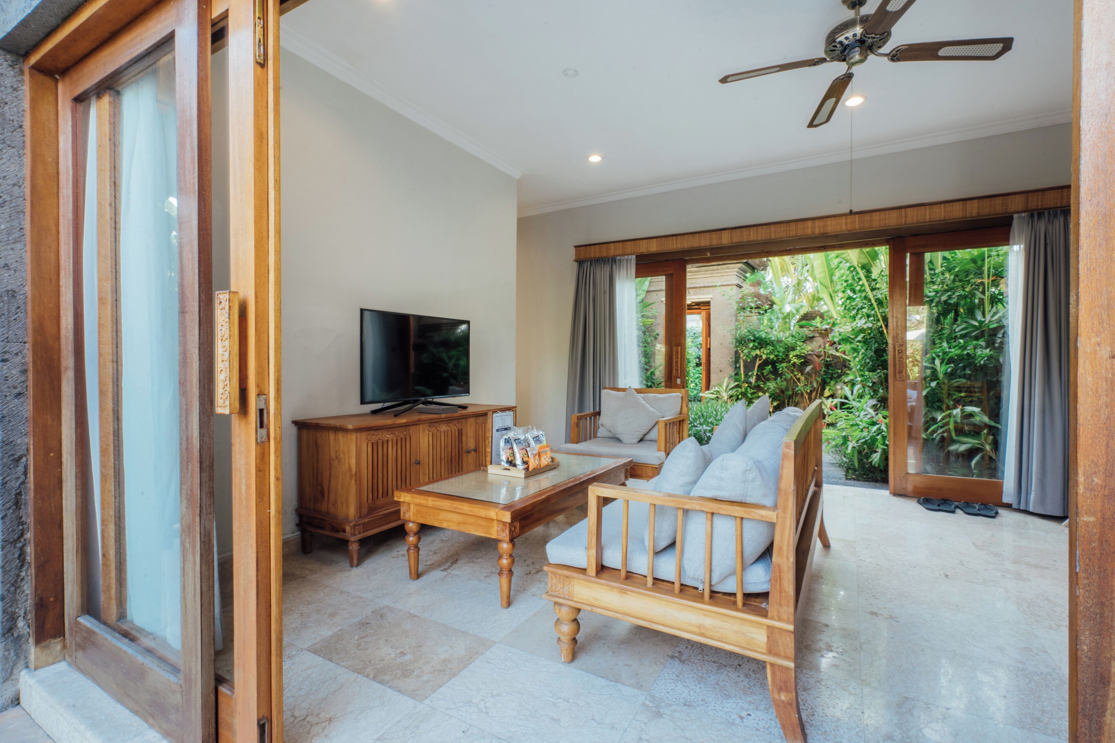 Una sala de estar con un sofá de madera, una mesa de centro, un televisor y un ventanal corredizo que da a un jardín.