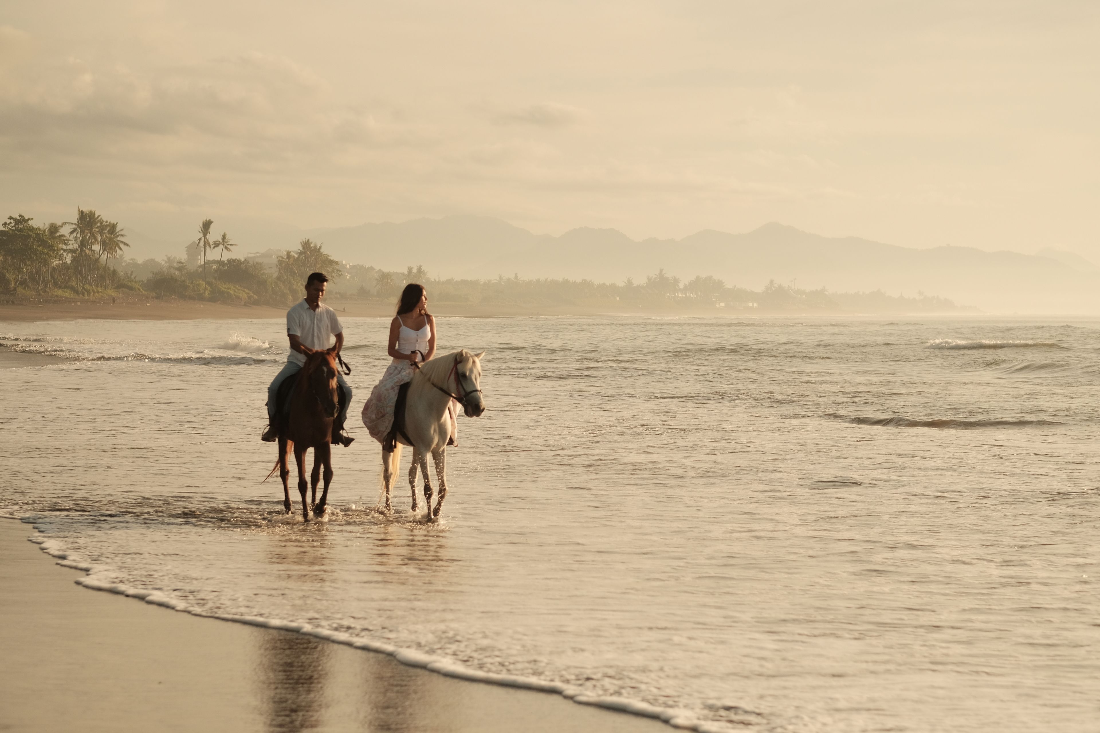 Two people riding horses along a beach with a view of the ocean and distant mountains.