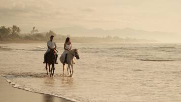 Two people riding horses along a beach with a view of the ocean and distant mountains.