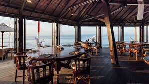 An open-plan dining area with wooden tables and chairs, large windows offering ocean views, and a high ceiling with exposed beams. - Desa Swan Villas & SPA (Gianyar)