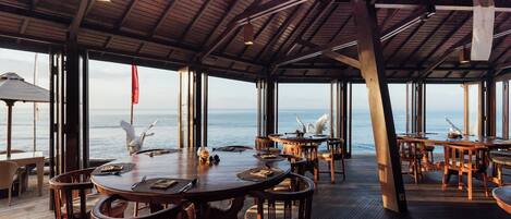Comedor de concepto abierto con mesas y sillas de madera, ventanas amplias con vistas al océano y un techo alto con vigas a la vista.