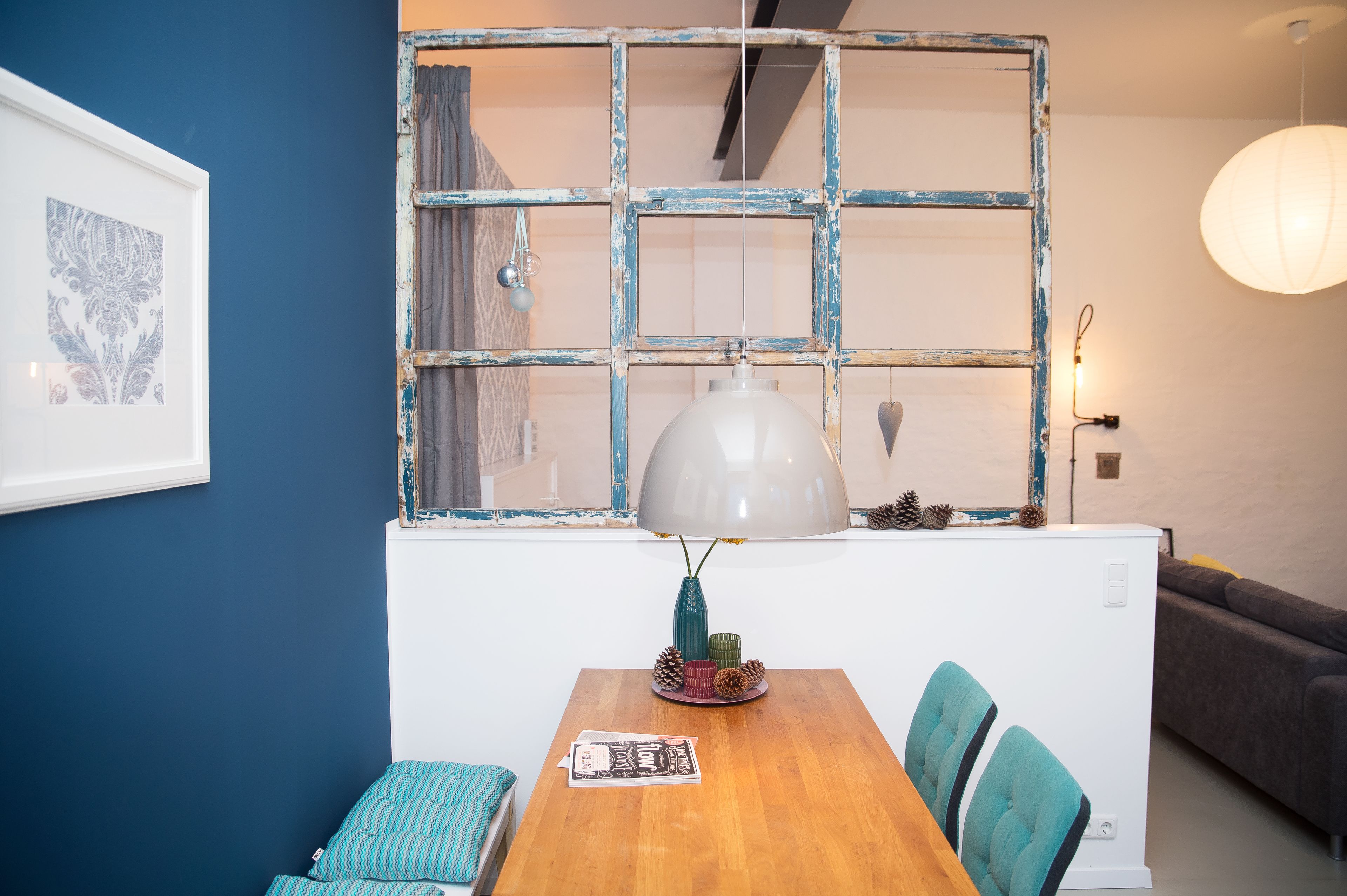 Un comedor con una mesa de madera, sillas color turquesa, una lámpara blanca y un marco de ventana decorativo.