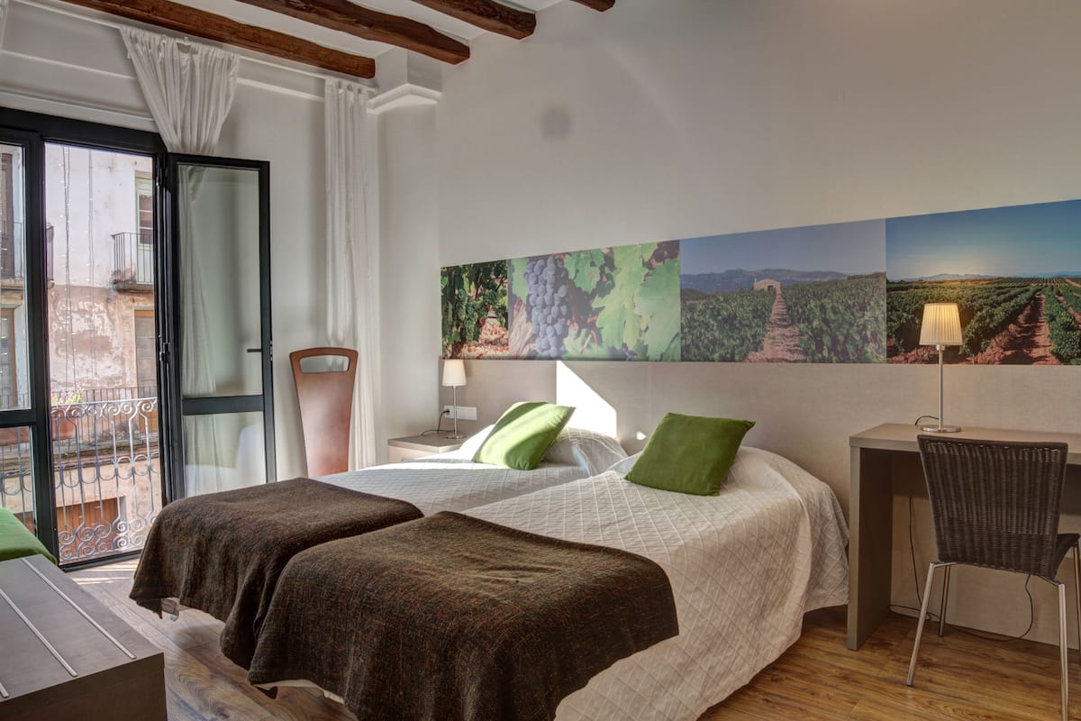 Habitación de hotel con dos camas, un escritorio, una silla y un ventanal con vistas a un viñedo.