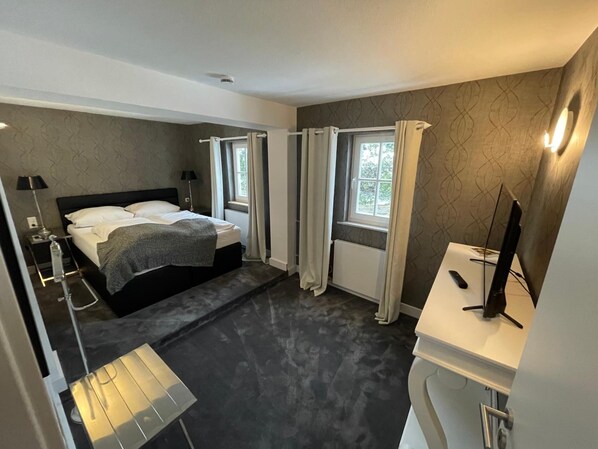 A modern bedroom with a bed, bedside table, lamp, and a view of the outdoors through a window. - Hotel Haus Roedgen (Wilnsdorf)