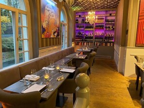 A restaurant with a long wooden table set for dining, a wine rack in the background, and a view of the outdoors. - Hotel Haus Roedgen (Wilnsdorf)