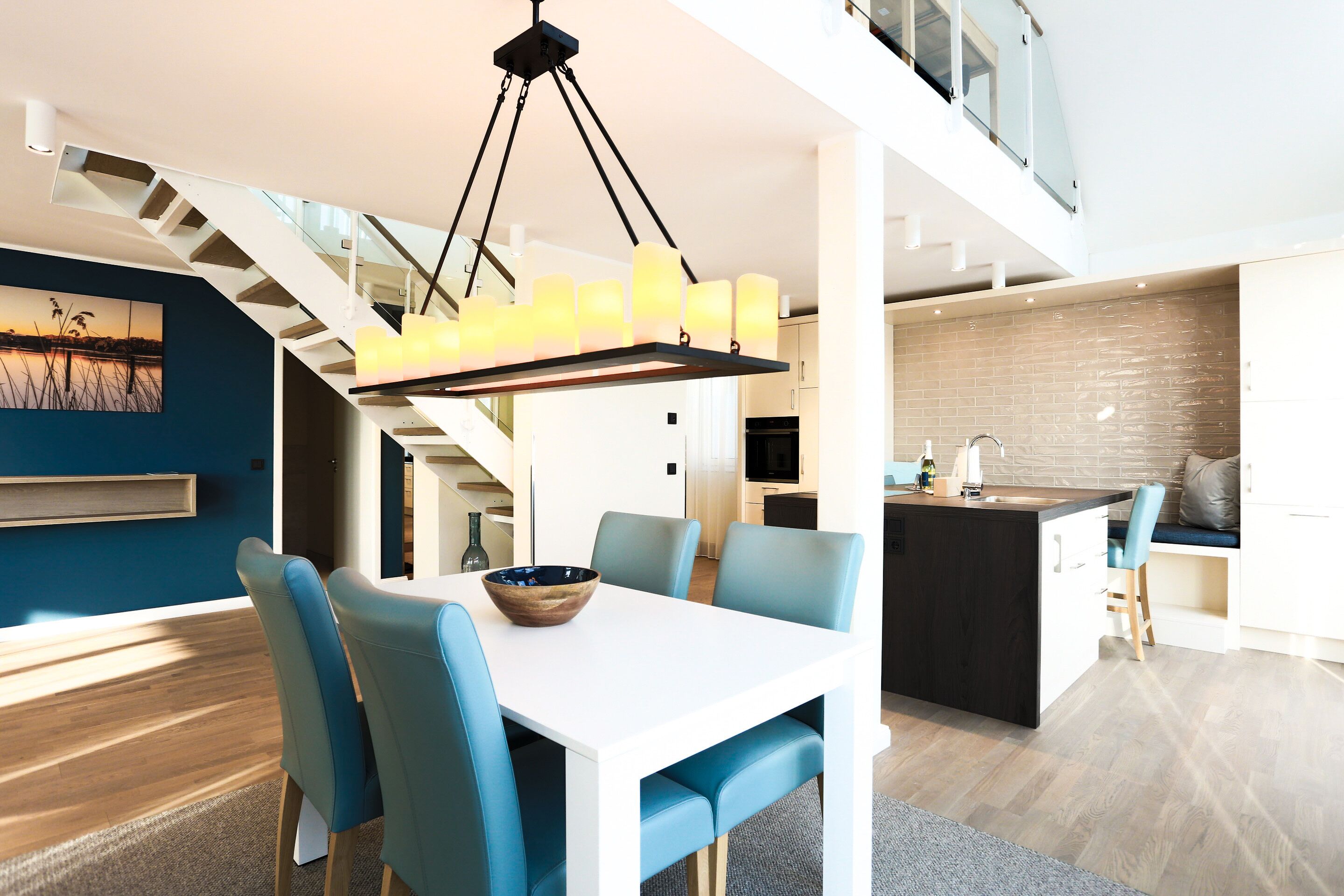 A modern kitchen with a central island, a dining area with blue chairs, and a staircase leading to an upper level.