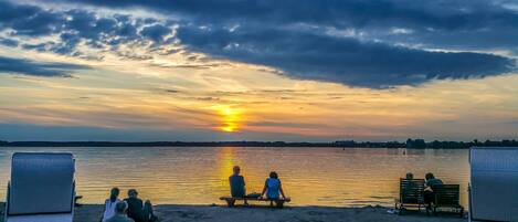 En strand med människor som sitter på bänkar och stolar och blickar ut över ett stilla vatten under solnedgången.
