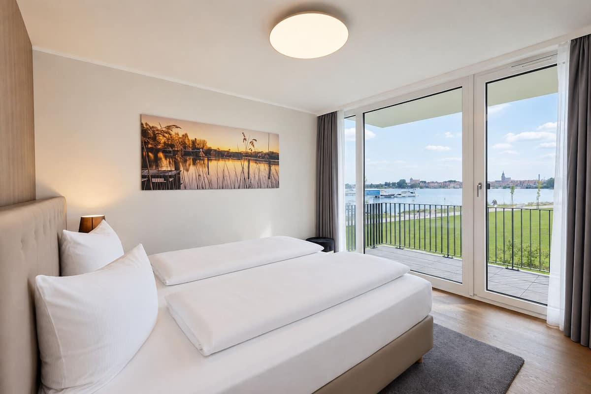 Quarto de hotel com uma cama grande, uma pintura do pôr do sol e vista para a água e para o horizonte urbano.