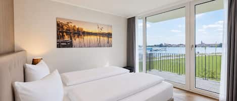 A hotel room with a large bed, a painting of a sunset, and a view of a body of water and cityscape.