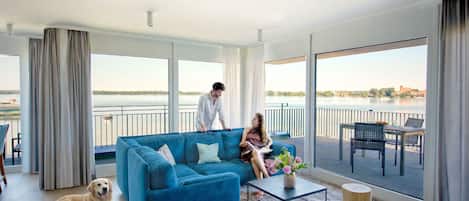 A spacious living area with a large blue sofa, a coffee table, and a dog. There is a balcony with a view of a body of water and a distant building.