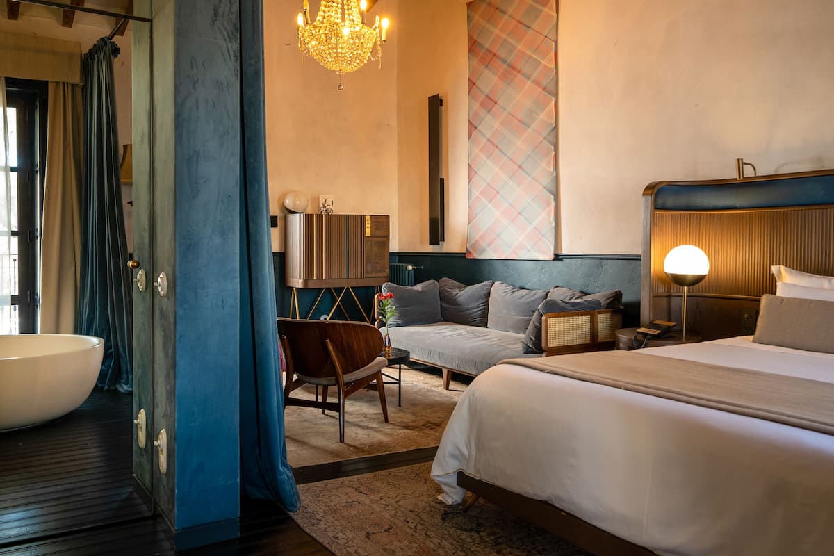 Une chambre d’hôtel avec un grand lit, un coin salon comprenant un fauteuil et une petite table, une grande fenêtre avec des rideaux et un lustre.