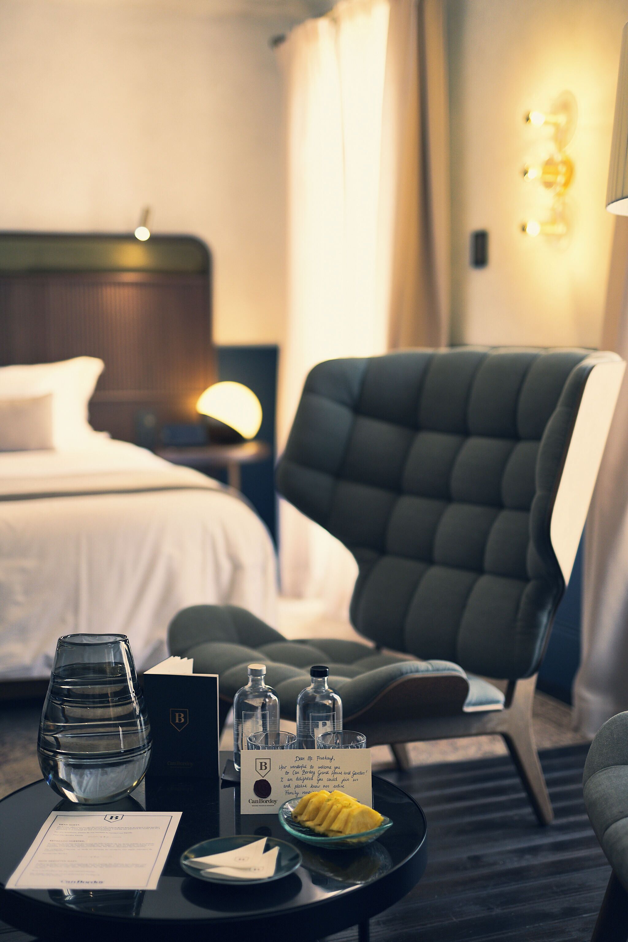 A hotel room with a bed, a chair, a table with a glass, a bottle, a menu, and a bowl of fruit.