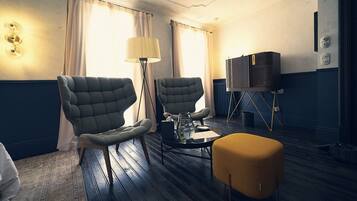 Une chambre d’hôtel avec deux fauteuils rembourrés, une petite table ronde, un lampadaire, un téléviseur posé sur un meuble et une grande fenêtre avec des rideaux.