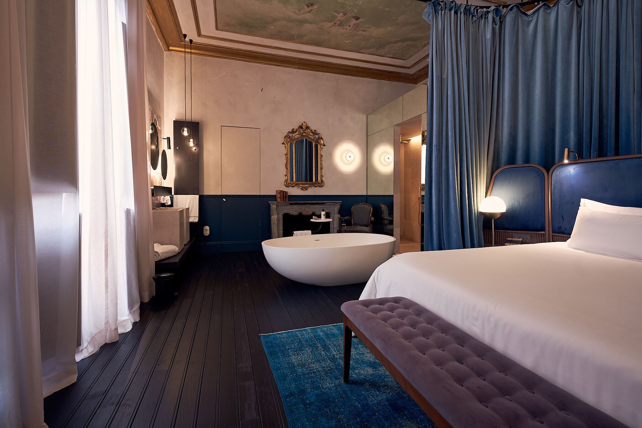 A hotel room with a large bed, a bathtub, a vanity with a mirror, and a ceiling with a fresco.