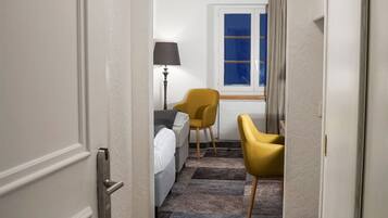 A hotel hallway with a checkered carpet, a bed with white linens, a yellow armchair, and a lamp.