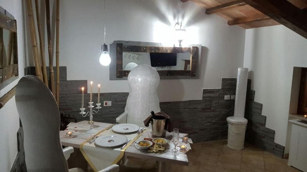 A dining area with a table set for two, a modern chandelier, a stone accent wall, and a television mounted above the fireplace.