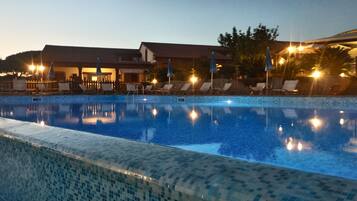 A swimming pool with lounge chairs and umbrellas, surrounded by buildings with a tiled roof.