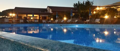 A swimming pool with lounge chairs and umbrellas, surrounded by buildings with a tiled roof.
