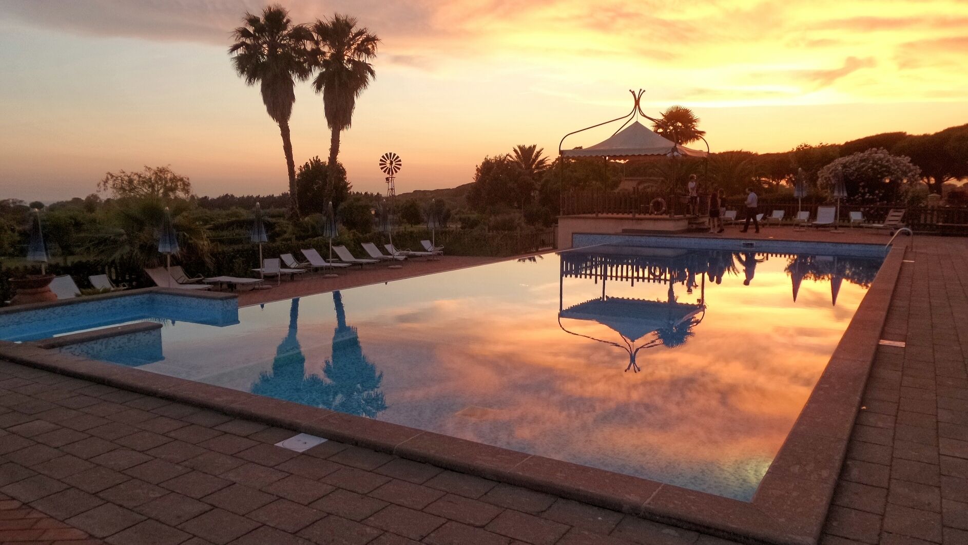 A swimming pool with lounge chairs and palm trees, reflecting the sunset.