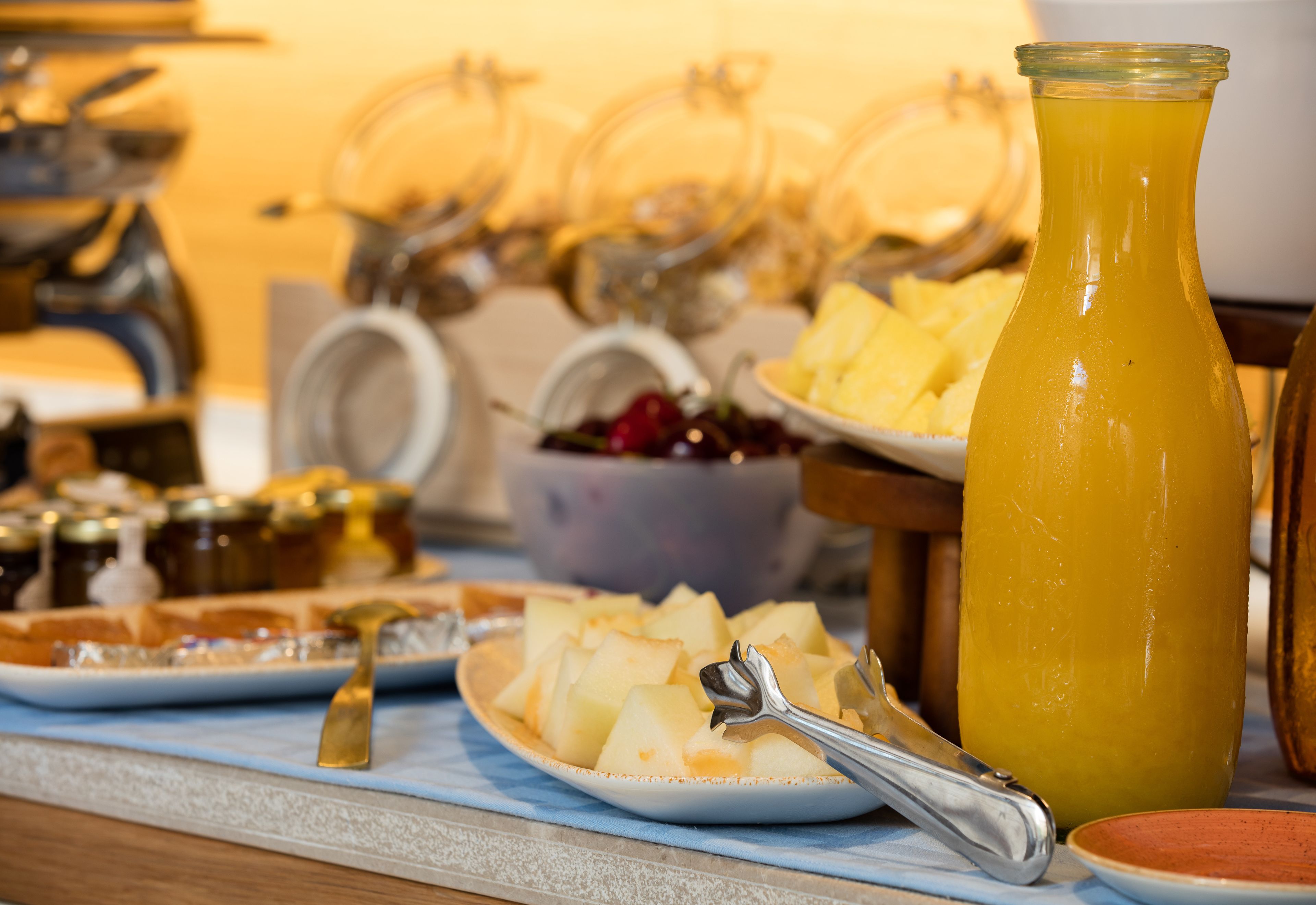 Une table de buffet proposant divers plats, notamment une assiette de pommes de terre en tranches, une bouteille de jus d’orange et plusieurs récipients contenant des condiments.