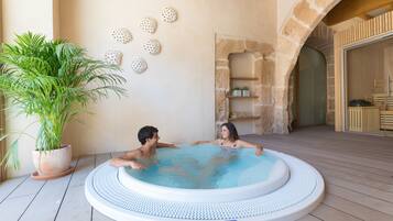 Un jacuzzi con dos personas, rodeado de una planta en maceta y conchas decorativas en la pared.