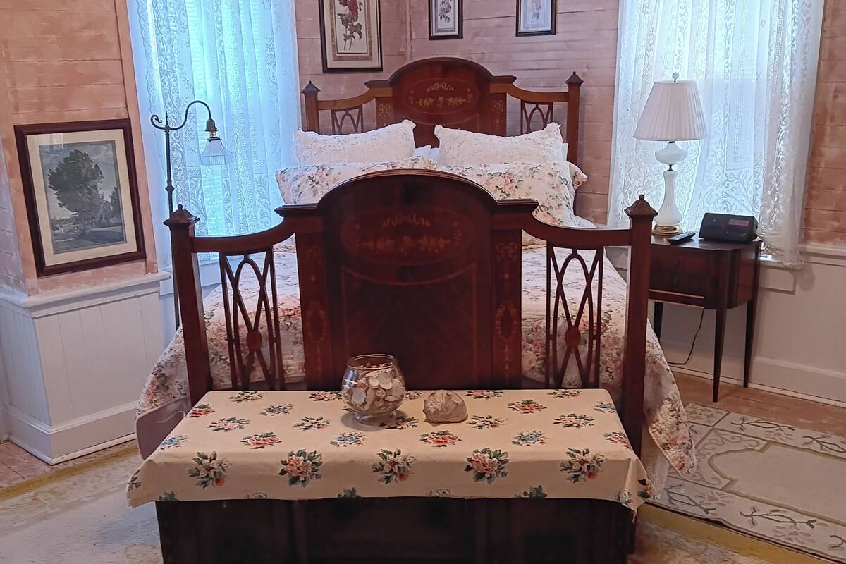 A bedroom with a wooden bed, floral bedspread, framed pictures on the wall, and a bedside table with a lamp.