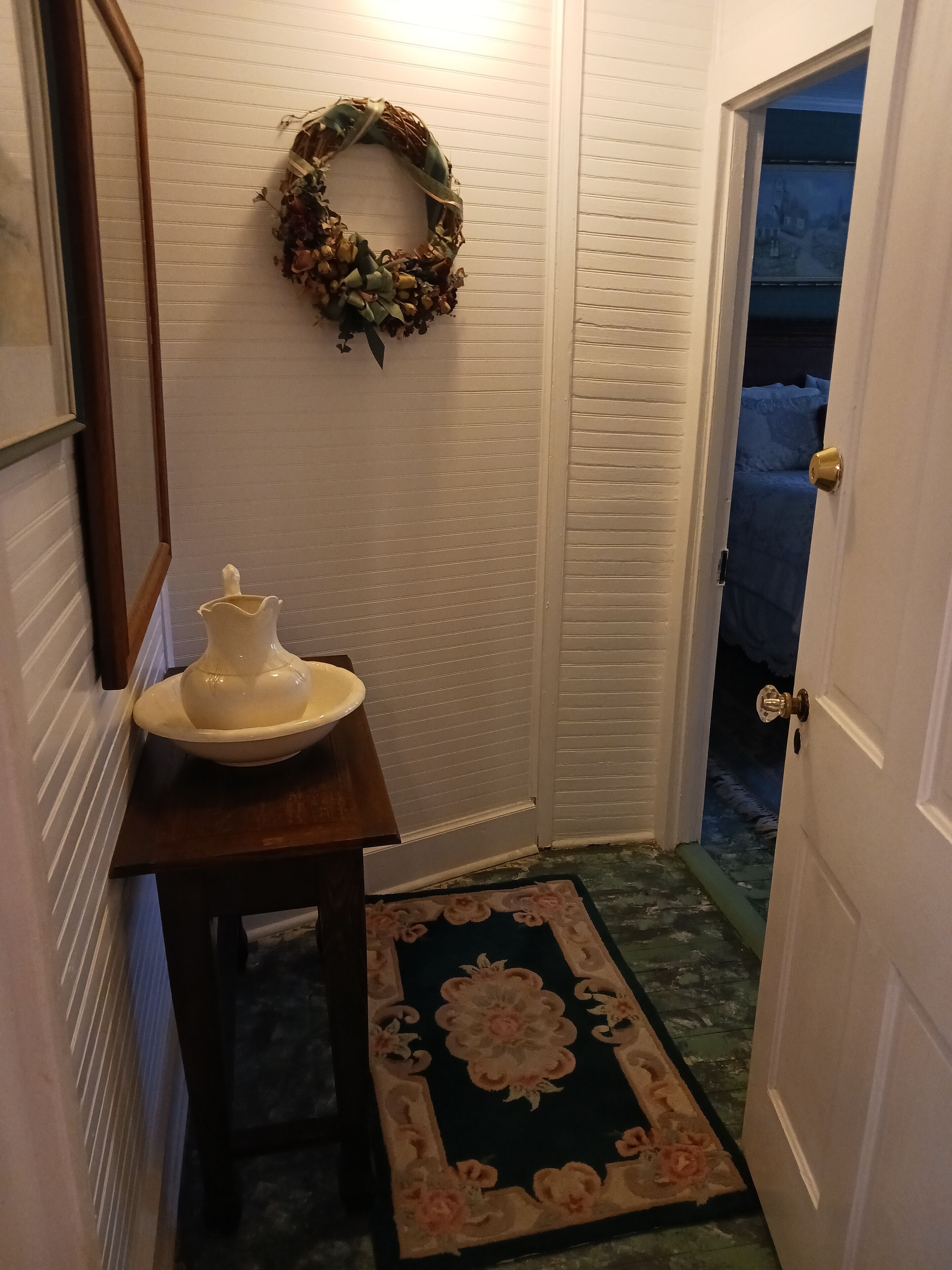 A hallway with a white door, a wooden table with a ceramic pot, a floral rug, and a wreath on the wall.