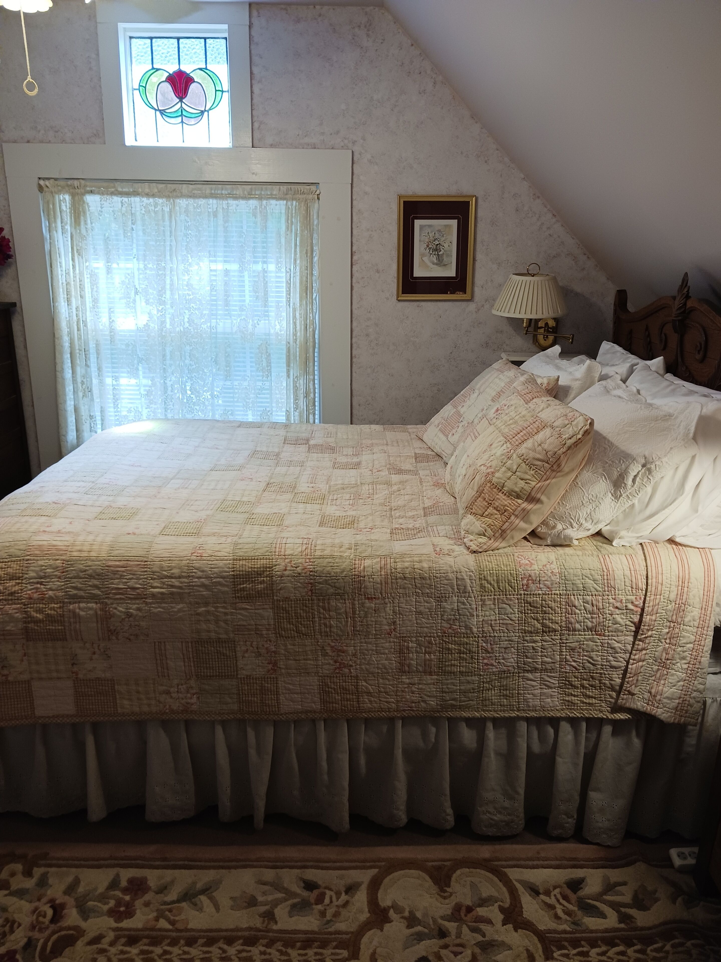 Une chambre à coucher avec un grand lit, une fenêtre ornée de vitraux, un tableau encadré au mur et une lampe de chevet.