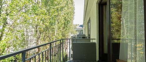A balcony with a view of trees, a railing, and a window with sheer curtains.