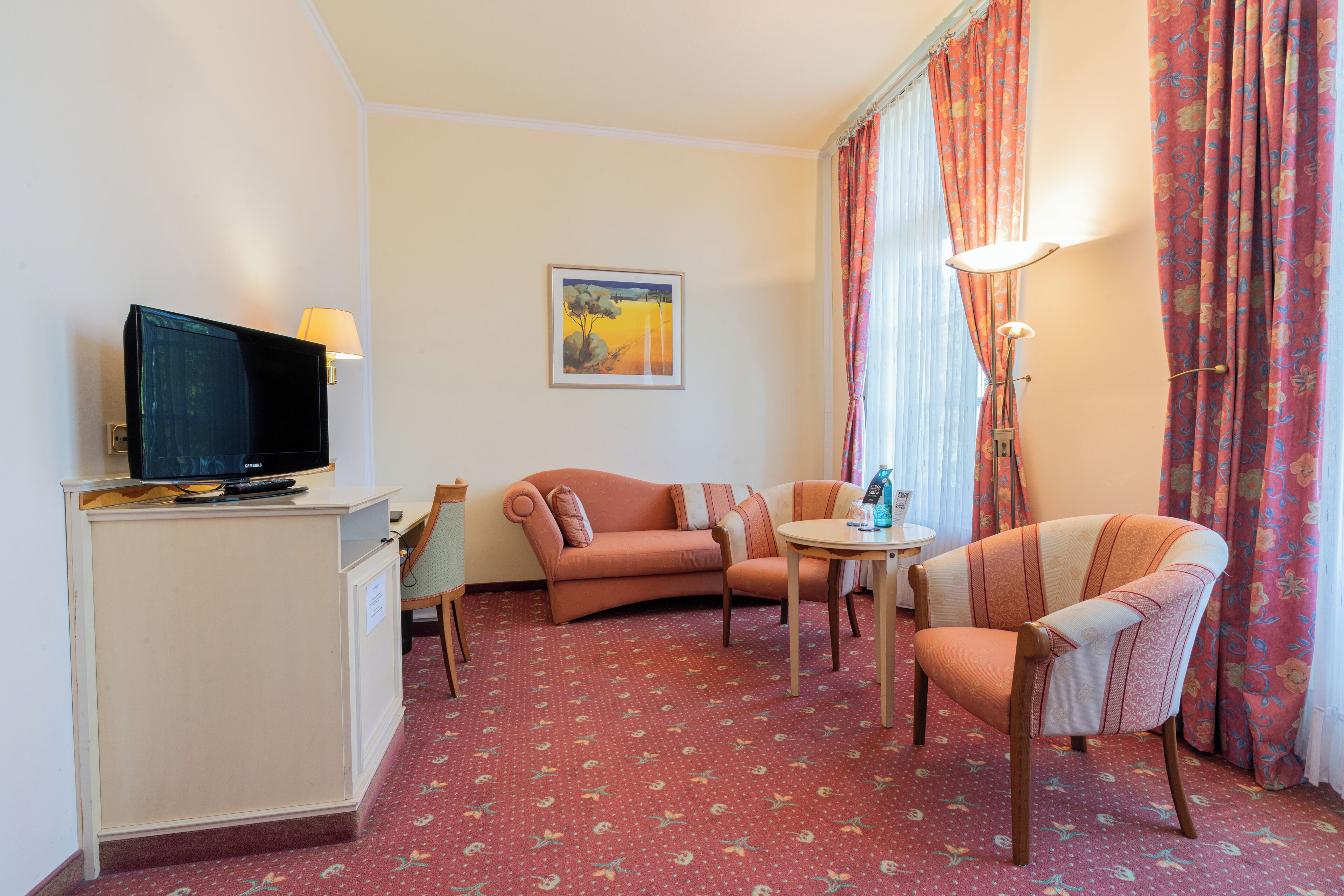 A hotel room with a television, a sofa, a chair, a small table, and a lamp.