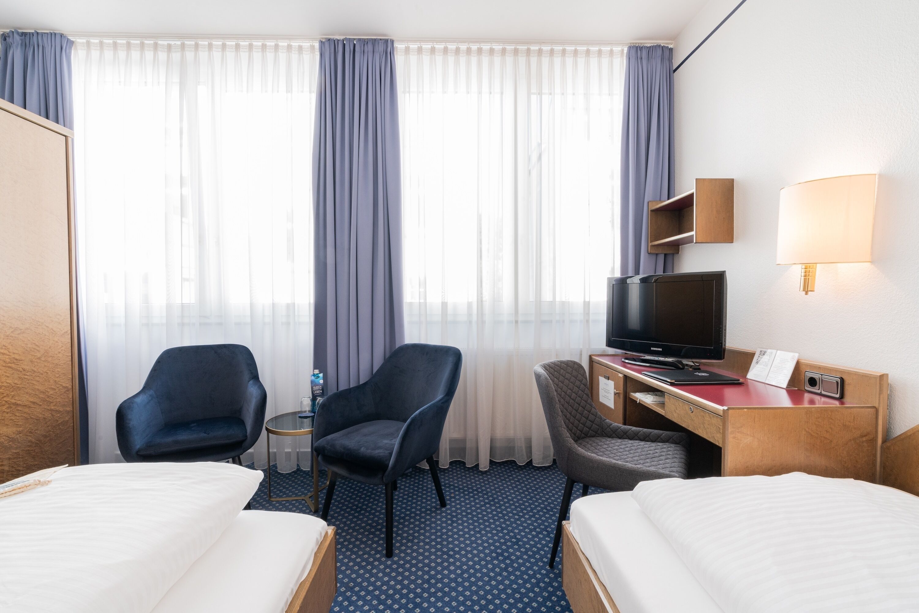 Habitación de hotel con dos camas, un escritorio con televisor, dos sillas, una mesita con una botella y una ventana con cortinas.
