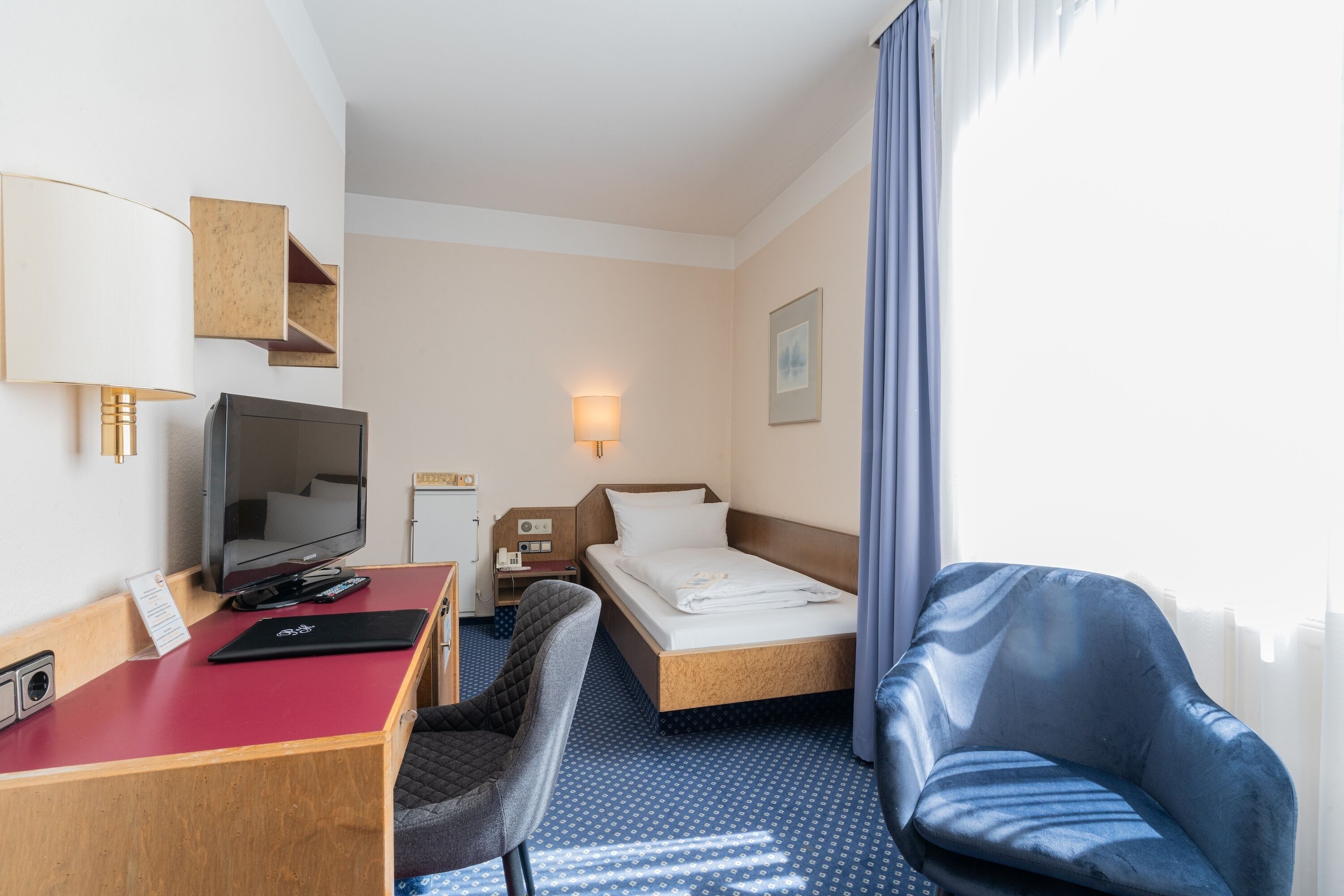A hotel room with a desk, television, bed, and a blue armchair.