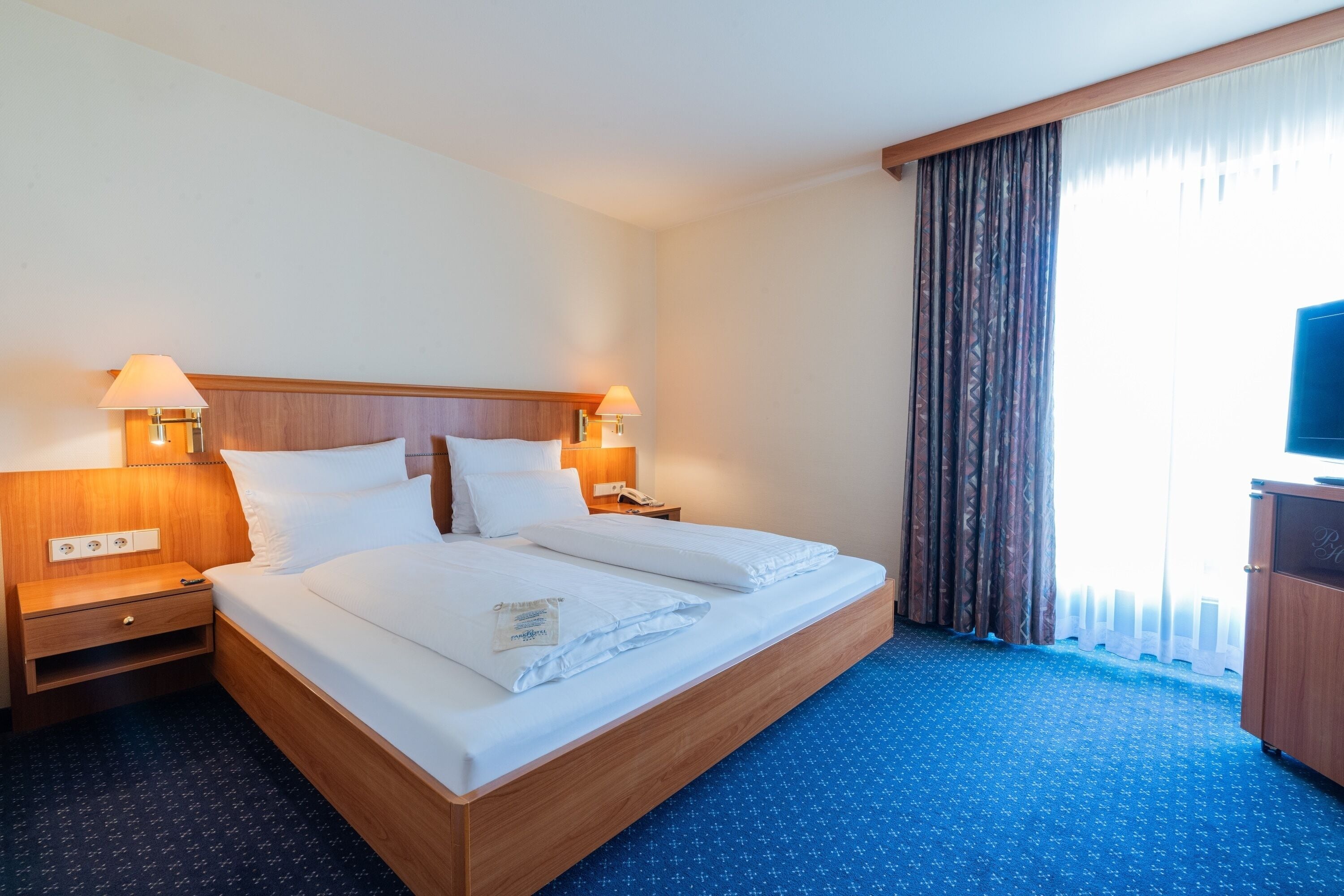 Ein Hotelzimmer mit einem hölzernen Bett, zwei Nachttischlampen, einem Nachttisch, einem Fernseher und einem Fenster mit Vorhängen.