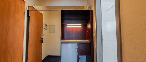 A narrow hallway with a wooden door, a small refrigerator, and a cabinet with a light.