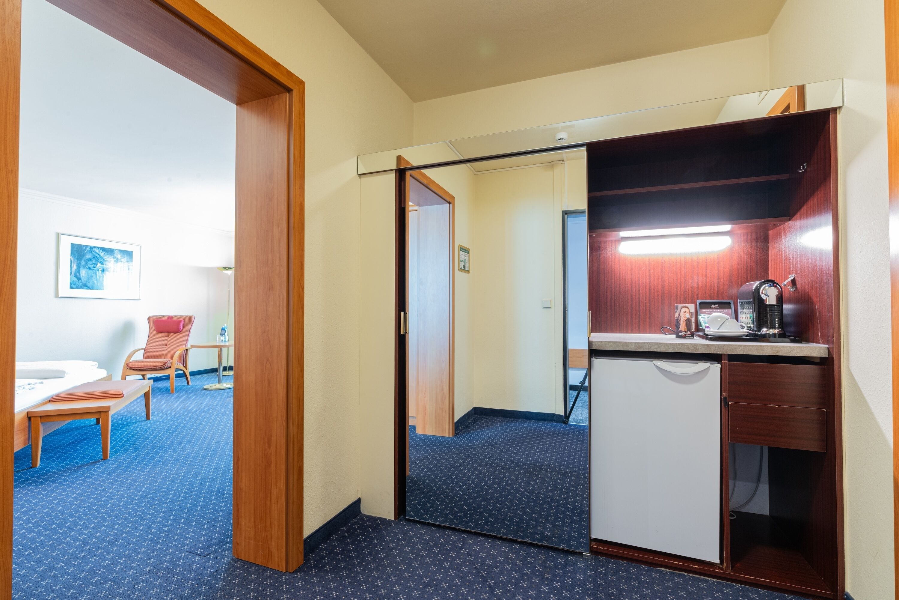 A hotel room with a kitchenette, a small seating area with a chair and table, and a door leading to another room.