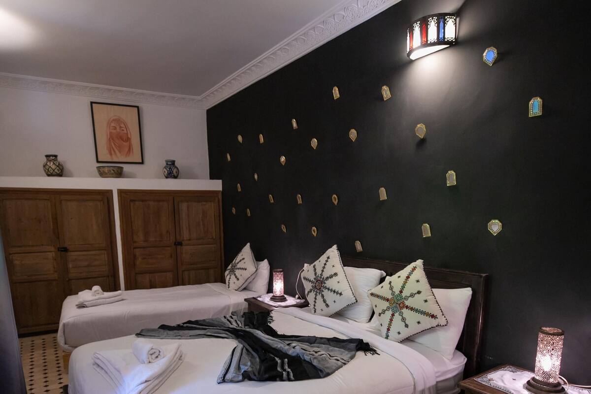 A bedroom with two beds, a nightstand with a lamp, patterned rugs, and a dark wall with decorative elements.