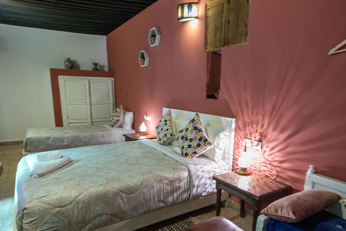 A bedroom with two beds, a nightstand, a lamp, a wardrobe, and a patterned rug.