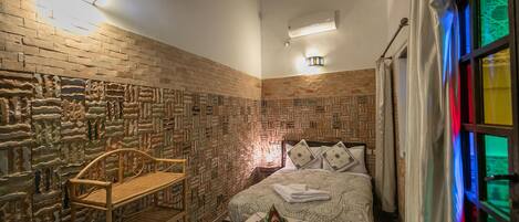 A room with a brick-patterned wall, a bed with patterned bedding, a small table with a decorative item, and a bench.