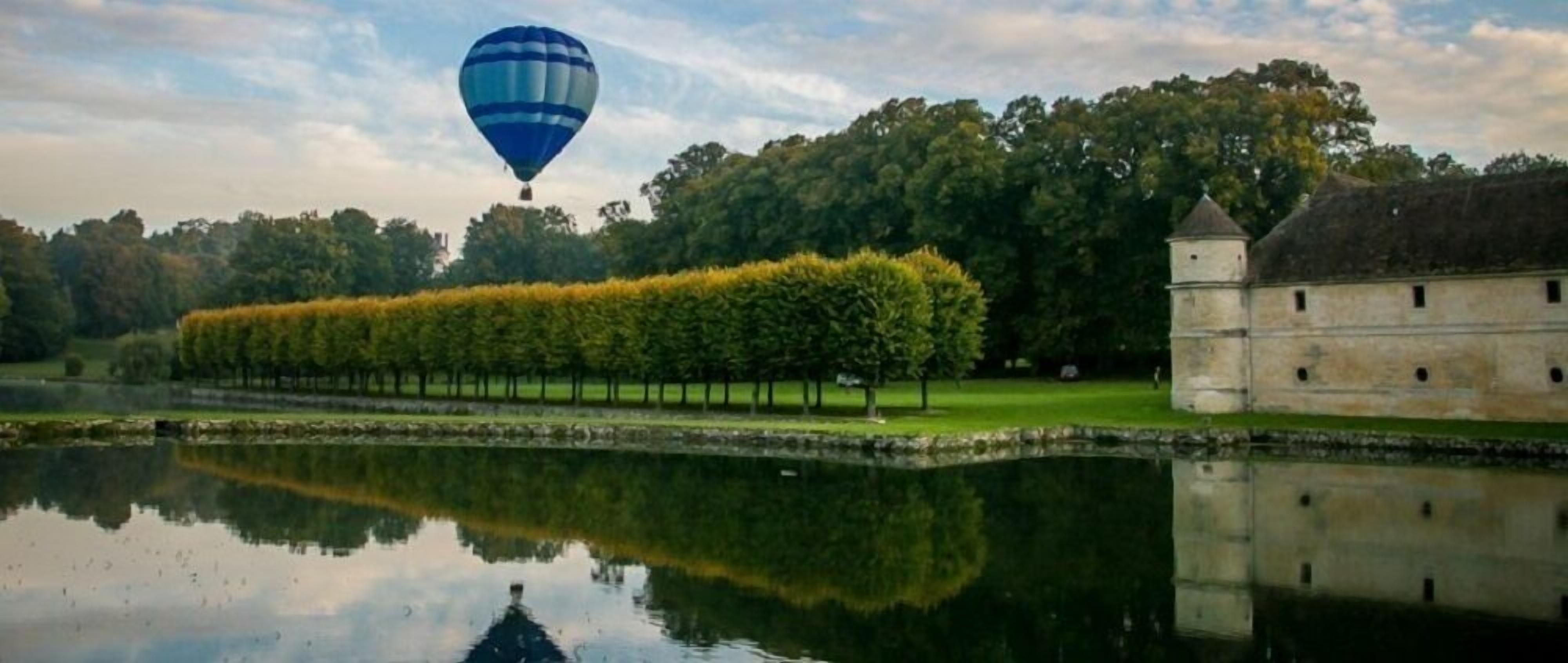 Une montgolfière survolant un lac, avec une rangée d’arbres et un bâtiment historique en arrière-plan.