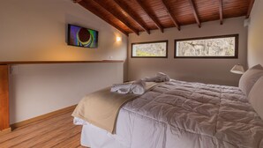 A bedroom with a wooden ceiling, a bed with a beige blanket, a wall-mounted TV, and two windows with natural light. - Andino Aparts - El Chalten (El Chalten)