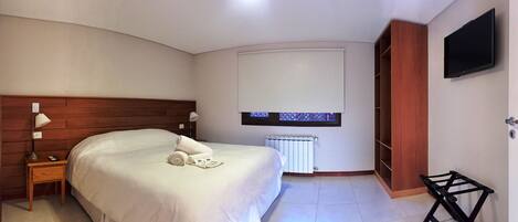 Habitación de hotel con cabecera de madera, cama con ropa blanca, mesita de noche con lámpara, televisor montado en la pared y radiador.