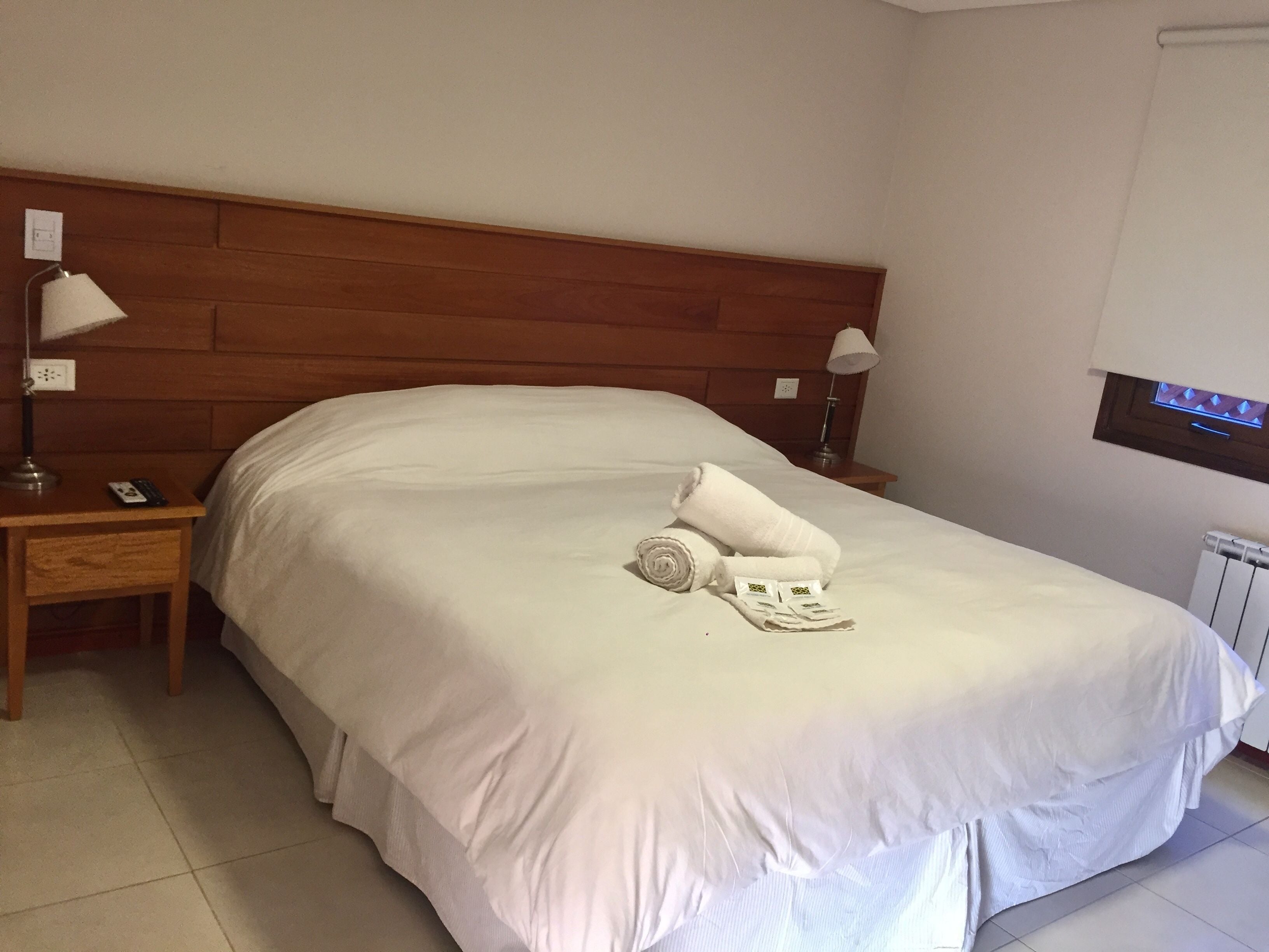 Una cama bien hecha con sábanas blancas, cabecera de madera y mesita de noche con lámpara y teléfono.