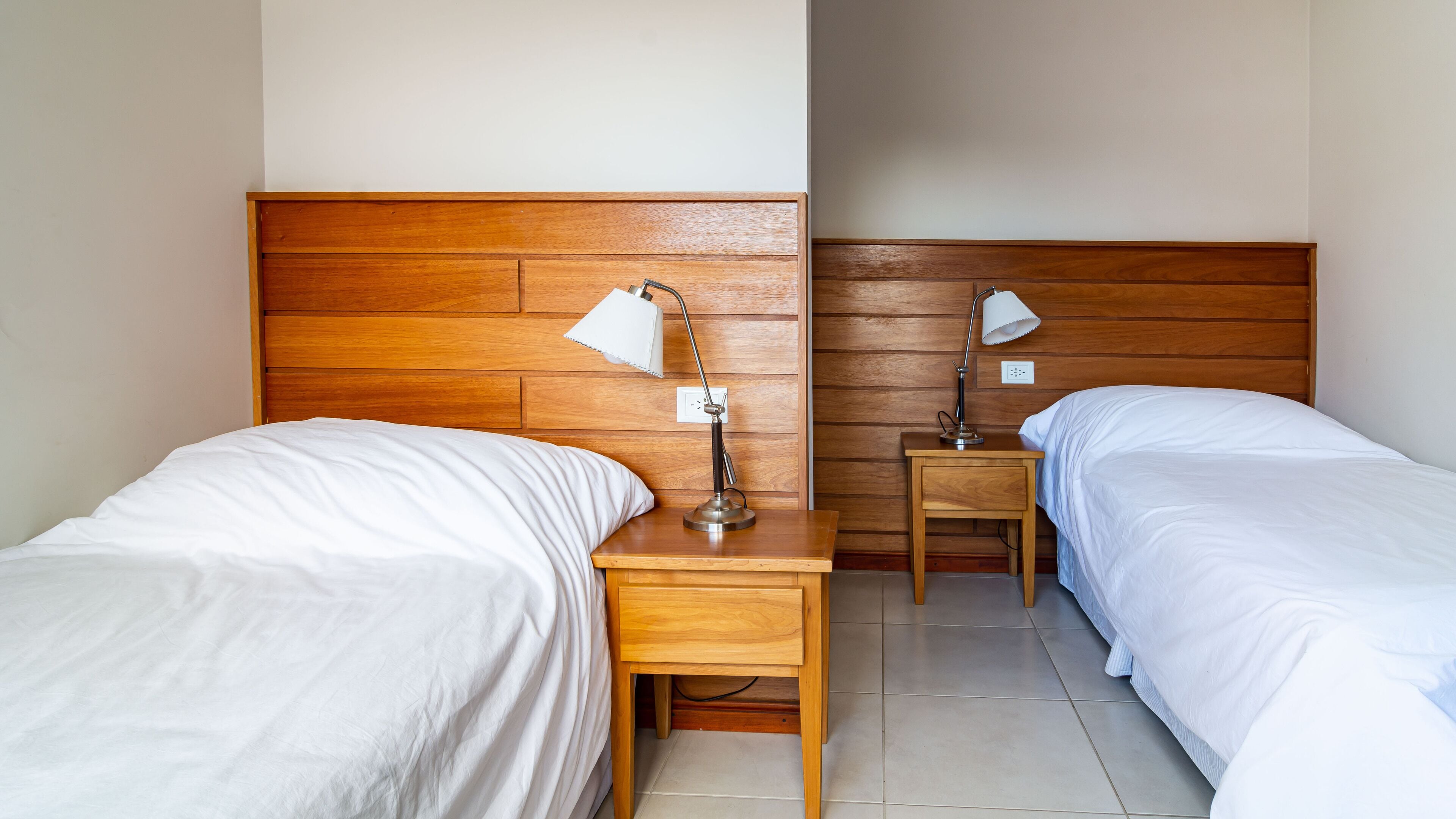 Habitación de hotel con dos camas individuales, cabeceros de madera, una mesita de noche con lámpara y un enchufe en la pared.