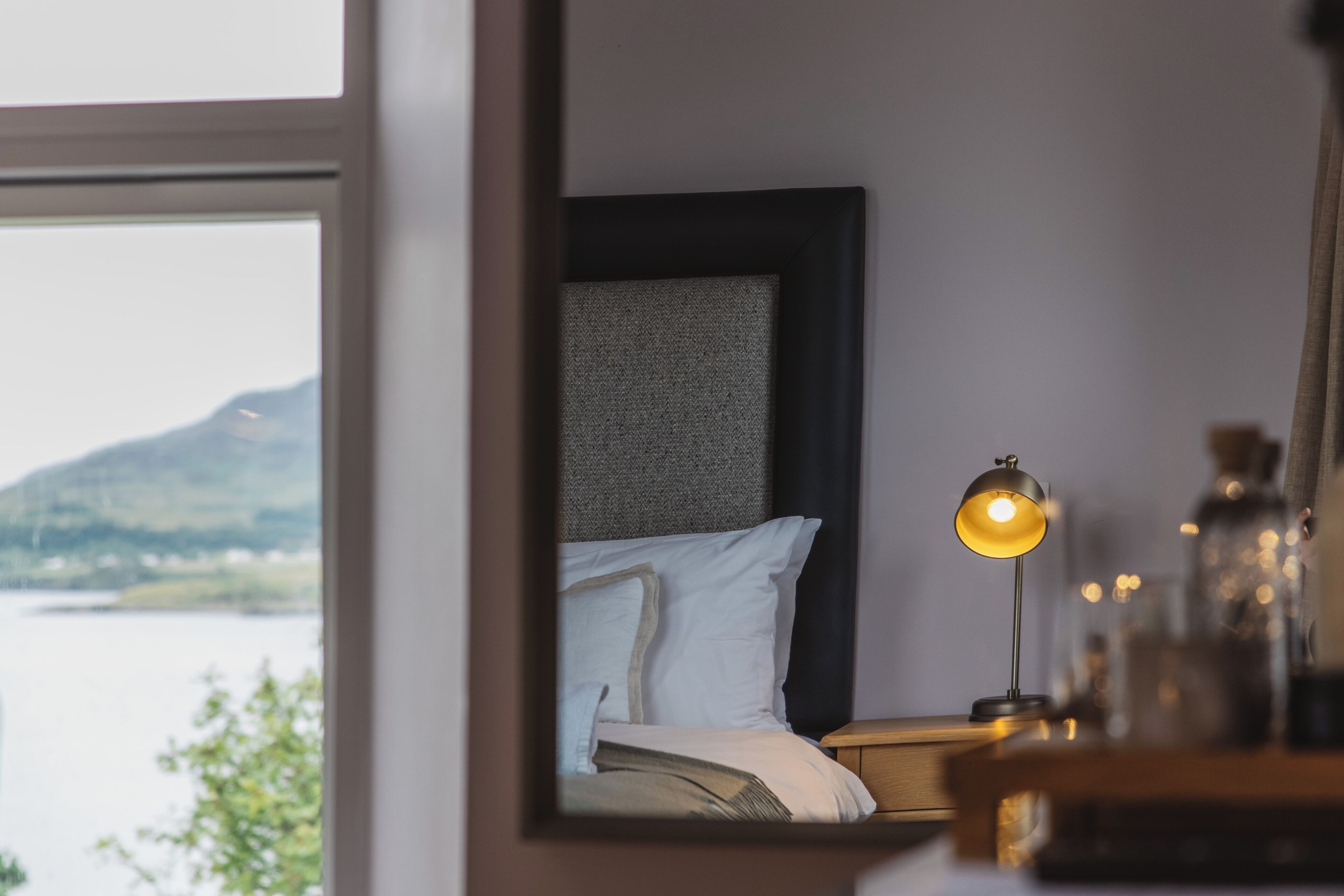 Un dormitorio con cama, mesita de noche, lámpara y vistas a un cuerpo de agua y colinas.